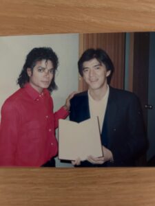 Michael Jackson con camisa roja posando junto a un hombre que sostiene un documento abierto en un entorno interior.