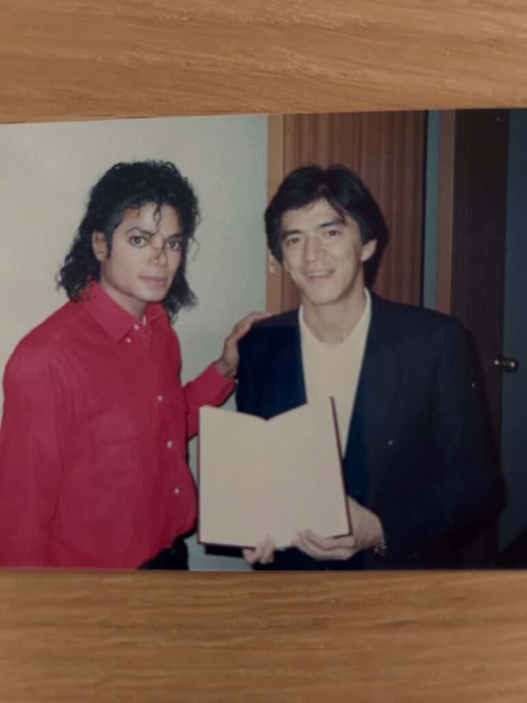 Michael Jackson con camisa roja posa junto a un hombre que sostiene un documento abierto.