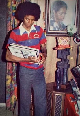 Michael Jackson joven con peinado afro, vistiendo una camisa de rayas horizontales rojas y azules, sosteniendo un álbum musical en una habitación decorada.